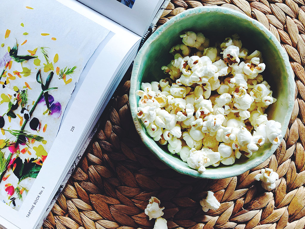 a a bowl with popcorn lays on top of a handwoven placemat next to an open book
